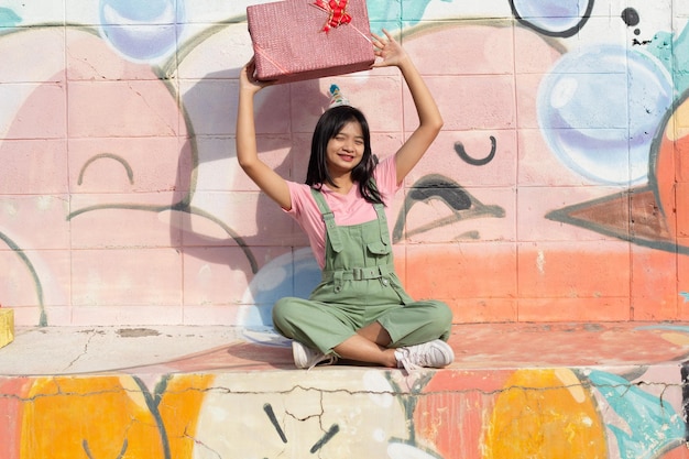 A garota alegre usando chapéu de festa com caixa de presente sentado no fundo colorido