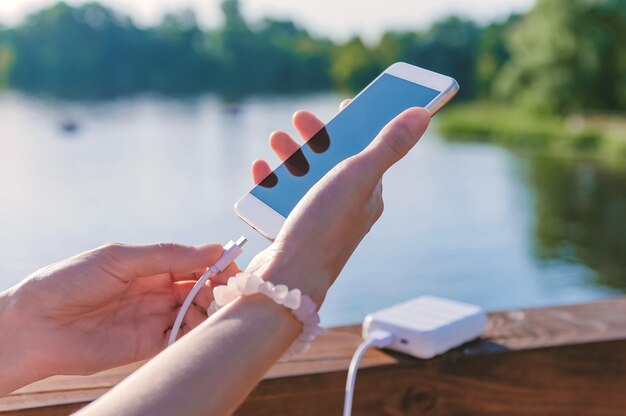 A garota carrega um smartphone branco do Power Bank em um píer de madeira no contexto da água.