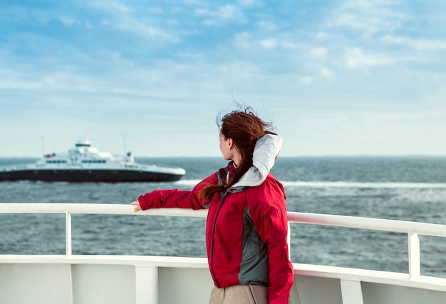 A garota com uma jaqueta vermelha na balsa olha para o mar, onde o transatlântico flutua