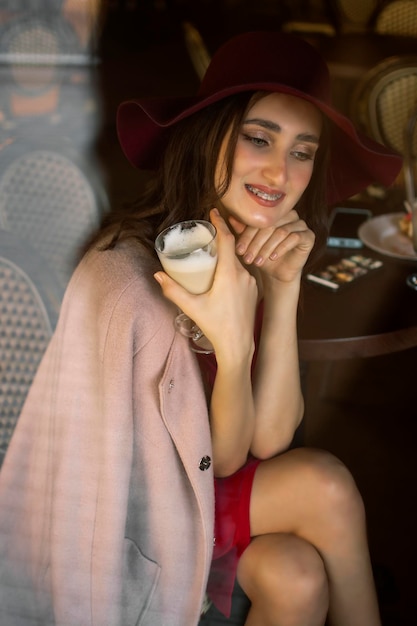 A garota come uma rosquinha e bebe café. Uma jovem morena de vestido vermelho come uma rosquinha rosa em um café no café da manhã.