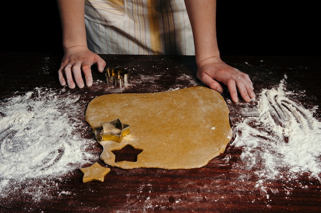 A garota de avental corta biscoitos em forma de massa em forma de estrelas em uma mesa de madeira. Fechar-se.