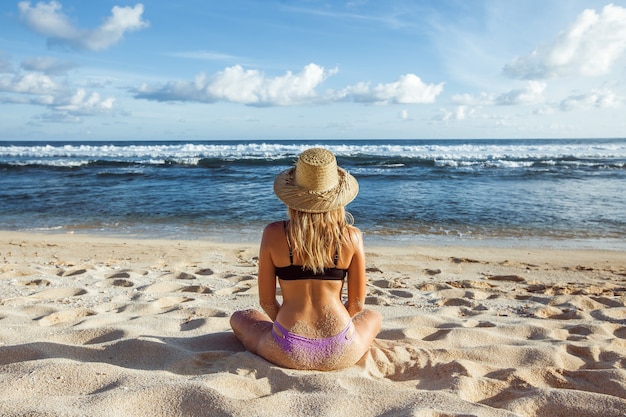 A garota de chapéu e maiô na praia se senta para a câmera e olha para o oceano