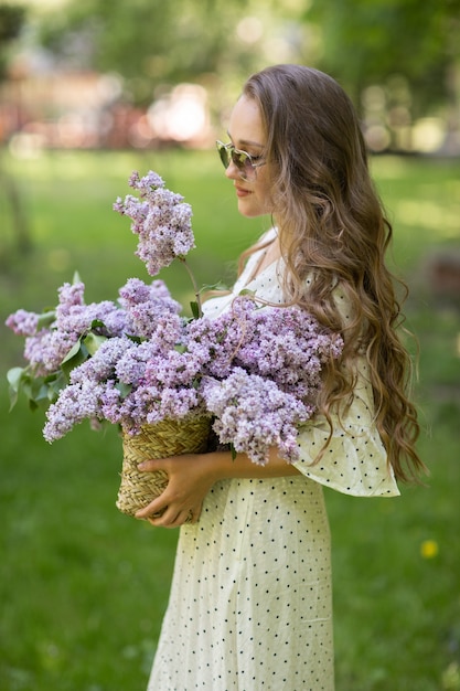 A garota de vestido branco e óculos escuros tem nas mãos uma cesta de vime com flores. Cesta com lilases. Menina e flores. Ande com uma cesta de lilases nas mãos. Florística