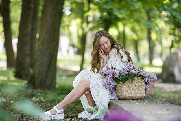 A garota de vestido branco tem nas mãos uma cesta de vime com flores e telefone. Cesta com lilases. Menina e flores. Sente-se com uma cesta de lilases nas mãos. Florística