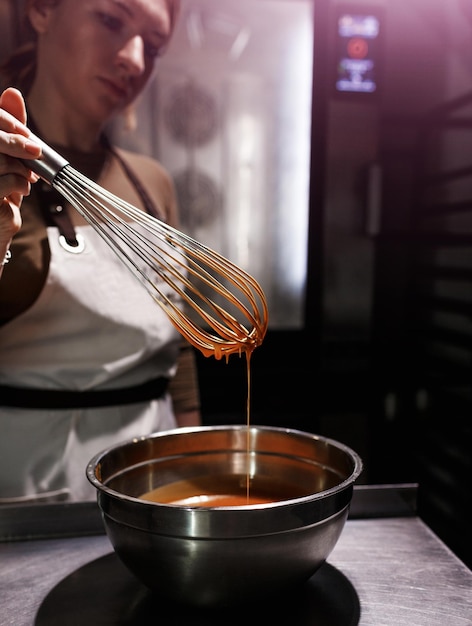 A garota do chef chicoteia o chocolate derretido em uma tigela com um close-up de uísque de arame velho na mão e nos pratos