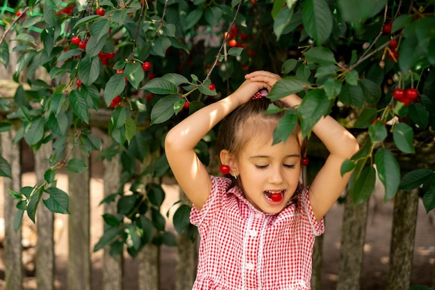 A garota está sentada debaixo de uma cerejeira e segurando uma baga vermelha na boca