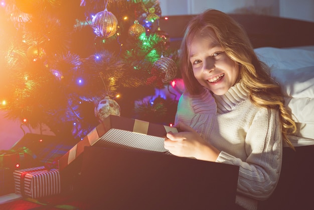 A garota feliz abre a caixa de presente perto da árvore de Natal. noite noite