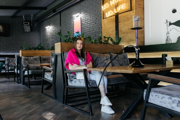 A garota fuma um narguilé Narguilé em um restaurante Mulher fuma um narguilé e se senta em uma cadeira A garota está sentada em um sofá em um café