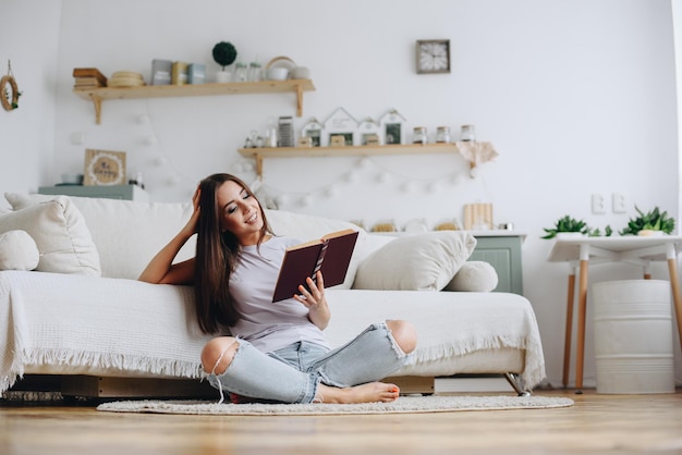 A garota lê um livro engraçado um romance em casa perto do sofá durante o dia Autodidata