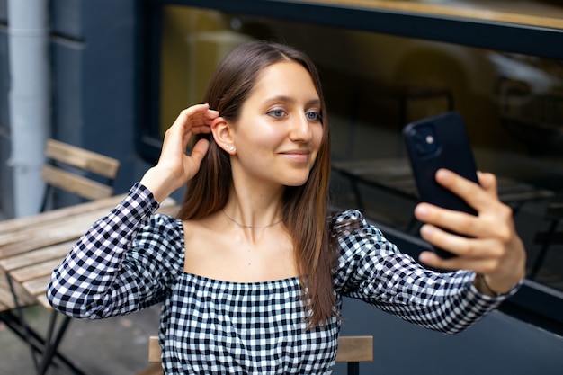 A garota moderna sorri e faz uma selfie ao telefone enquanto está sentada à mesa em um café ao ar livre