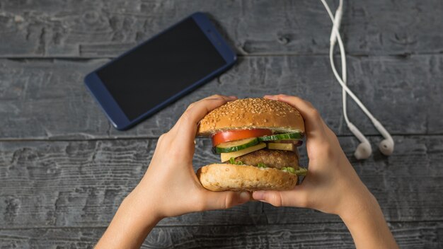 A garota segura um hambúrguer preparado sobre uma mesa com um telefone e fones de ouvido