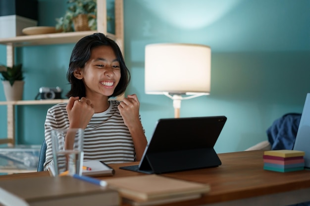 A garotinha asiática está estudando com sucesso online através da internet em um tablet digital enquanto está sentada em uma mesa em casa à noite. criança da ásia feliz e sorridente. conceito de aprendizagem online em casa