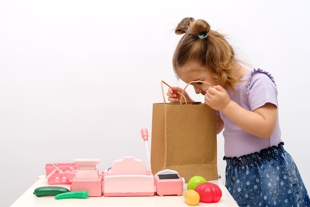 A garotinha brinca na loja olha para o saco de mantimentos brinquedo caixa registradora escala brinquedos vegetais e frutas na mesa pedidos on-line e entrega de alimentos