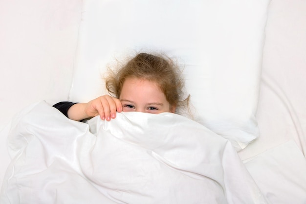 A garotinha na cama está coberta com um cobertor, apenas os olhos rindo são visíveis
