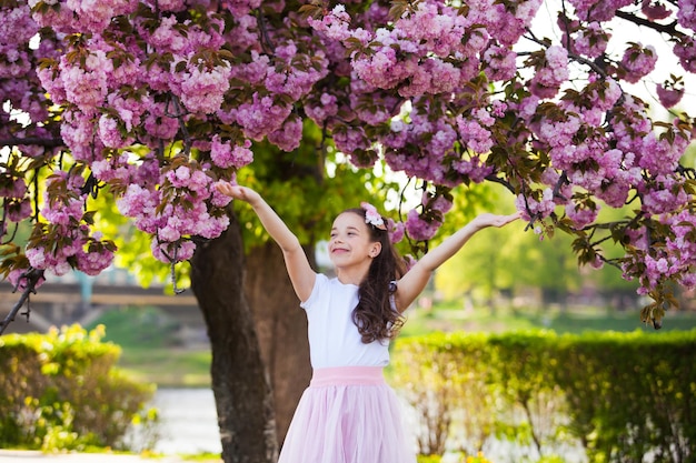 A garotinha sorridente está de pé sob uma sakura Ela alegremente espalha pétalas de flores