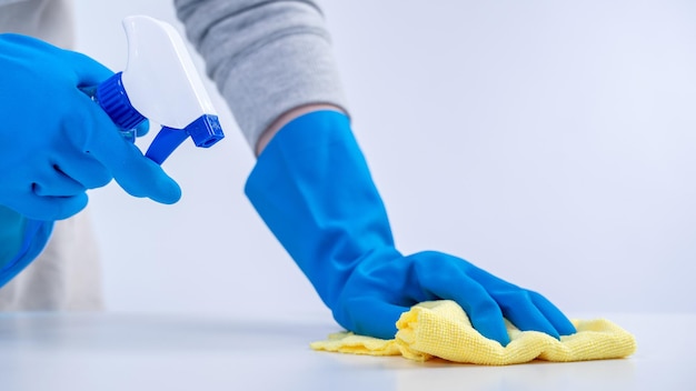 A governanta jovem no avental está limpando a superfície da mesa com luvas azuis, pano amarelo molhado, pulverizando garrafa, limpador de garrafa conceito de design closeup