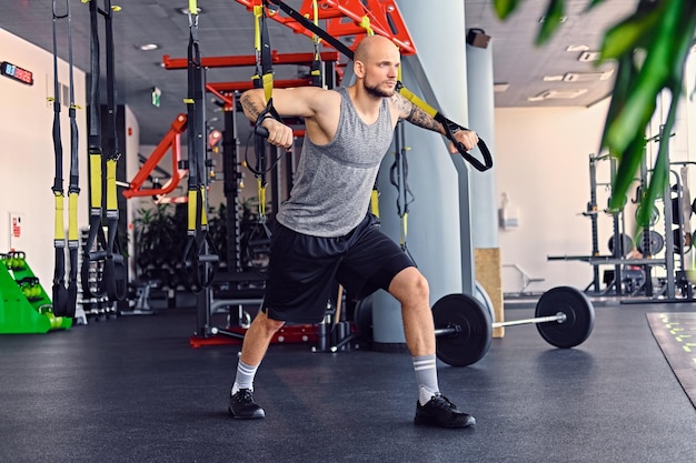 A imagem de corpo inteiro do homem de cabeça raspada e tatuado barbudo atlético fazendo exercícios de tiras trx em um clube de ginástica.