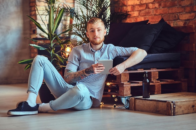 A imagem de corpo inteiro do homem elegante loiro vestido com uma camisa de lã e jeans usando um tablet PC em uma sala de estar.