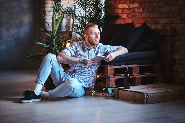 A imagem de corpo inteiro do homem elegante loiro vestido com uma camisa de lã e jeans usando um tablet PC em uma sala de estar.