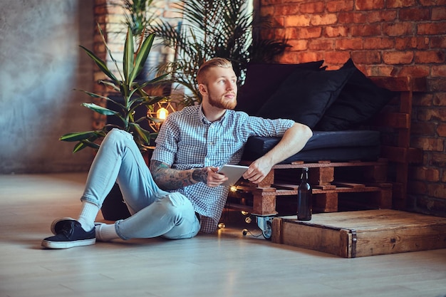 A imagem de corpo inteiro do homem elegante loiro vestido com uma camisa de lã e jeans usando um tablet PC em uma sala de estar.