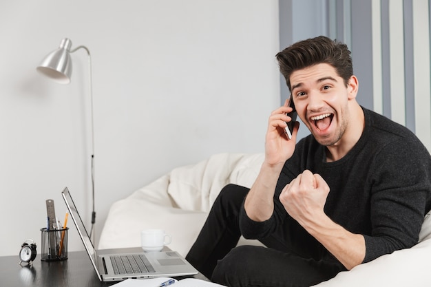 A imagem de um jovem bonito feliz em casa dentro de casa usando o computador laptop faz o gesto de vencedor falando pelo telefone celular.