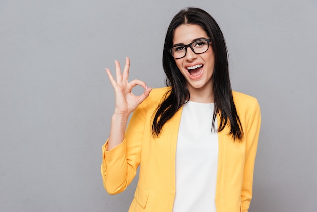 A imagem de uma jovem alegre usando óculos e vestida com uma jaqueta amarela faz o gesto de Ok sobre a superfície cinza. Olhe para a frente.