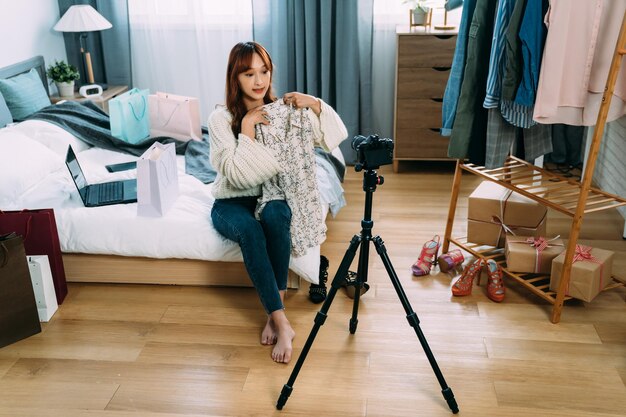 A influenciadora de beleza asiática sentada descalço na cama está apresentando e desembalando uma nova blusa na frente da câmera em transmissão ao vivo em casa com piso de madeira.