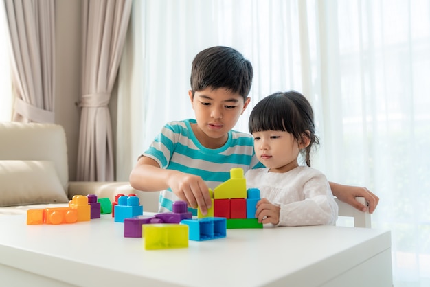 A irmã e o irmão bonitos asiáticos jogam com um designer de blocos de brinquedo em cima da mesa na sala de estar em casa.