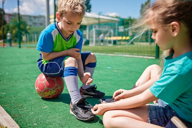 A irmã mais velha amarra os cadarços das botas ao irmão durante um jogo no campo de futebol.