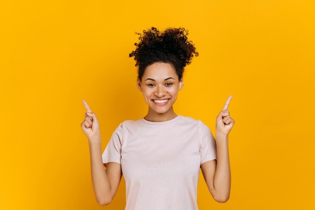 A jovem afro-americana amigável positiva de cabelos encaracolados olha para a câmera e aponta os dedos para cima no espaço vazio da cópia fica no fundo de cor laranja isolado sorrindo alegremente