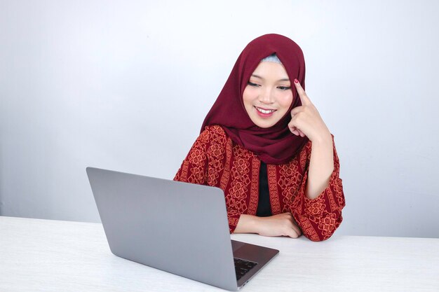 A jovem asiática islâmica está sorrindo com um gesto de pensamento olhando para o espaço em branco na frente do laptop