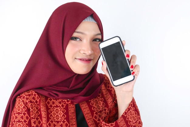 A jovem asiática islâmica está sorrindo mostrando o smartphone em pé sobre fundo branco