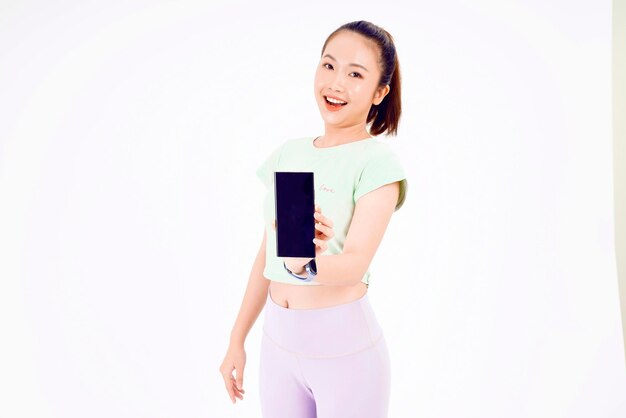 A jovem asiática mostra a tela vazia do smartphone com sorrisos de expressão positiva amplamente vestidos com roupas casuais, sentindo felicidade no fundo azul Celular com tela branca na mão feminina