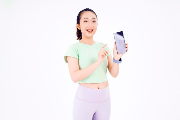 A jovem asiática mostra a tela vazia do smartphone com sorrisos de expressão positiva amplamente vestidos com roupas casuais, sentindo felicidade no fundo azul Celular com tela branca na mão feminina