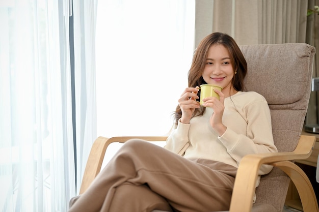 A jovem asiática relaxa na sala de estar sentada na poltrona confortável e bebe café quente