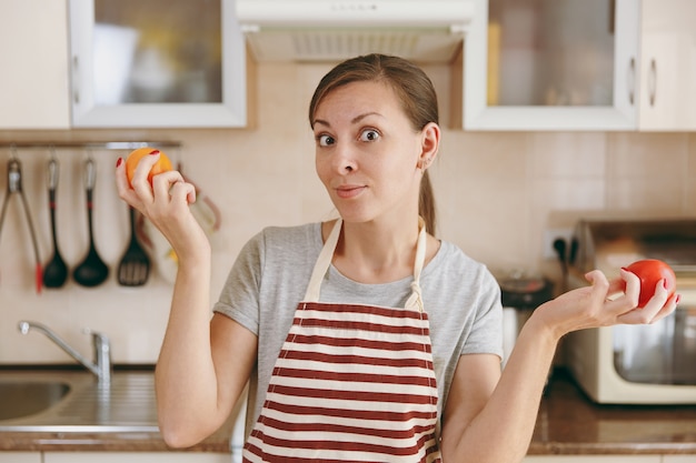 A jovem atraente e pensativa de avental decide escolher um tomate vermelho ou amarelo na cozinha. Conceito de dieta. Estilo de vida saudável. Cozinhando em casa. Preparar comida.