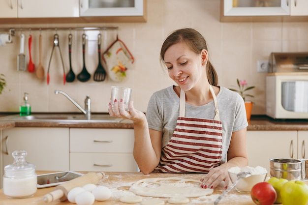 A jovem bela mulher feliz sentado à mesa com farinha e recorte um copo em círculos de massa para bolinhos na cozinha. cozinhando em casa. preparar comida.