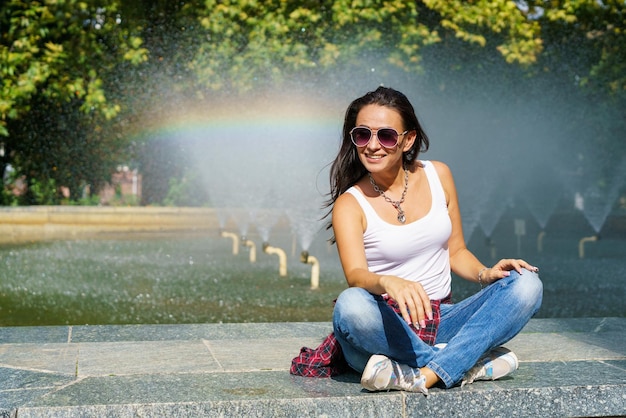 A jovem caucasiana sentada perto da fonte gosta de frescura da água no sol