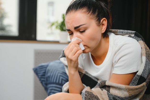 A jovem doente sente-se no sofá coberta com um cobertor congelando assoando o nariz escorrendo espirrando no tecido Fêmea com febre sintomas de gripe e gripe tosse em casa Doente alérgico com doença respiratória