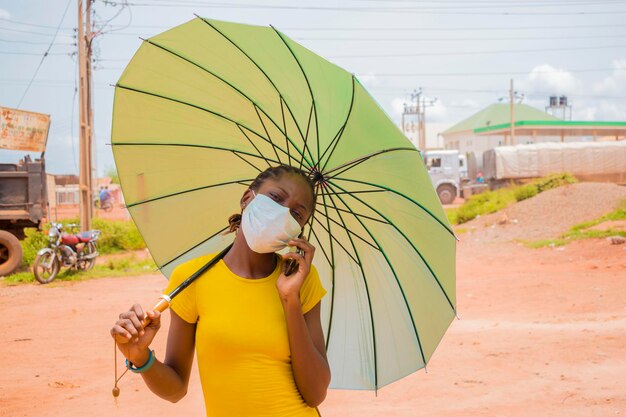A jovem e bela africana usando máscara facial preveniu-se do surto em sua sociedade em pé sob o sol, usando guarda-chuva como proteção e fazendo chamadas com o telefone