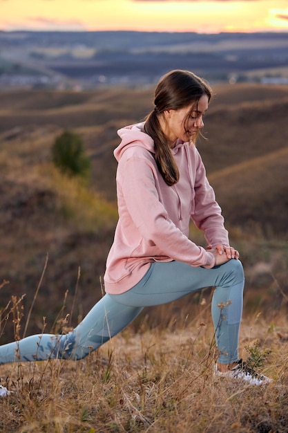 A jovem em forma entra para esportes no campo de verão ao pôr do sol. mulher saudável está envolvida em fitness, fora da cidade. Treino e aquecimento ao ar livre, respira ar fresco em campo, esticando as pernas