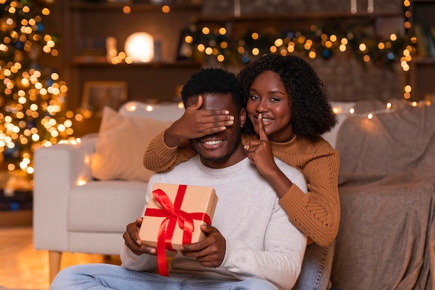 A jovem esposa afro-americana sorridente fecha os olhos para o marido mostra sinal de silêncio e dá caixa de presente