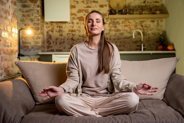 A jovem está descansando em casa meditando com os olhos fechados sentada no sofá da sala uma dona de casa morena na posição de lótus sorri