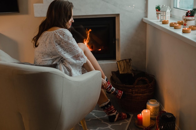 A jovem está sentada em uma sala aconchegante junto à lareira A garota está sentada em uma poltrona e veste meias quentes de natal