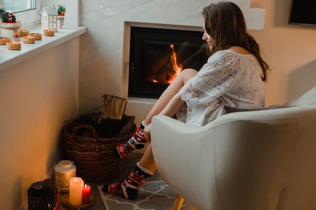 A jovem está sentada em uma sala aconchegante junto à lareira A garota está sentada em uma poltrona e veste meias quentes de natal