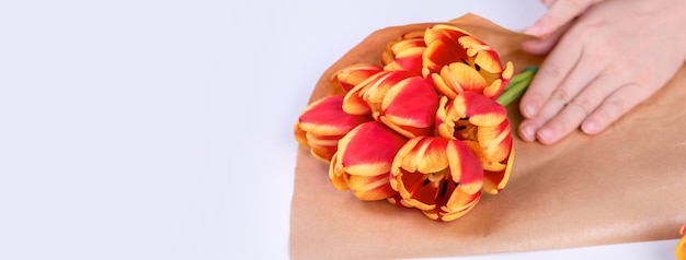 A jovem florista feminina está embrulhando um buquê de flores de tulipa de cor vermelha para o presente do dia das mães com laço de fita de gravata sobre um fundo de mesa branca close-up estilo de vida