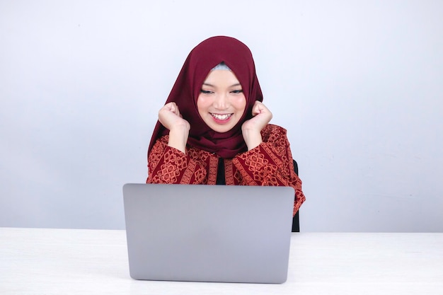A jovem islâmica asiática usando lenço na cabeça está chocada e animada com o que vê no laptop na mesa