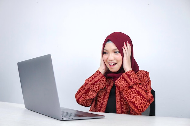 A jovem islâmica asiática usando lenço na cabeça está chocada e animada com o que vê no laptop na mesa