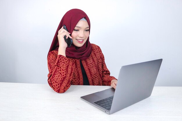 A jovem islâmica asiática usando lenço na cabeça está sorrindo e animada quando ela liga para o telefone e para a frente do laptop na mesa