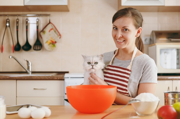 A jovem linda mulher feliz com um gato persa branco prepara massa para bolos com tablet na mesa da cozinha. cozinhando em casa. preparar comida.